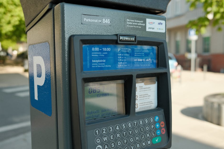 Close-up view of a parking payment machine, featuring a blue 'P' sign on the side, situated outdoors on a paved area. The machine has a digital screen displaying parking rates and timings, alongside a