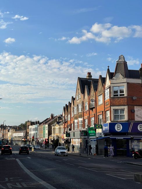 A street scene featuring a row of multi-storey residential buildings with brick facades and gabled roofs, situated along a busy commercial area on High Street Colliers Wood. In the foreground, there a