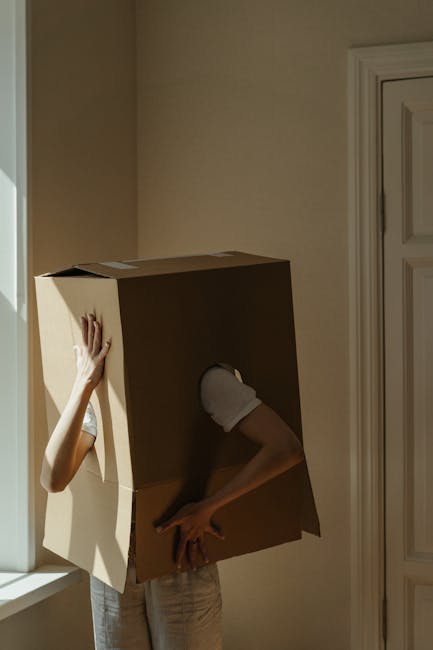 A young woman with brown hair seated on a wooden floor surrounded by large cardboard boxes during a home relocation process, inside a room with light blue textured walls and a grey stone accent wall.